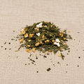 A small pile of loose-leaf herbal tea or matcha blend with dried green leaves, yellow seeds, and white flower petals, placed on a beige linen fabric background.
