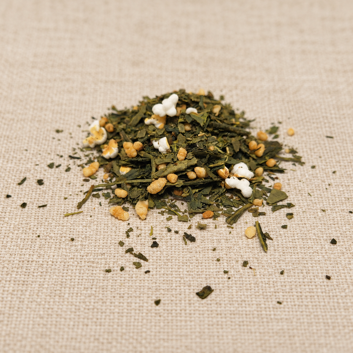 A small pile of loose-leaf herbal tea or matcha blend with dried green leaves, yellow seeds, and white flower petals, placed on a beige linen fabric background.